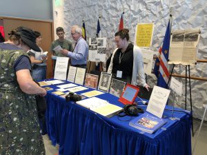 The PCCA exhibit features several posters on easels behind a blue table with seven more documents, two binders, and an IPad. A woman stands in front of the exhibit. Robert Newlen and Leslie Nellis stand behind the exhibit to answer questions.