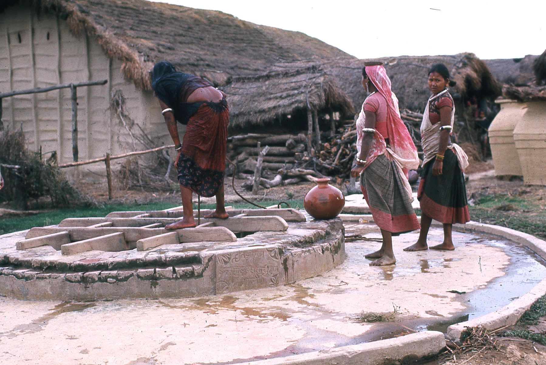 Three women collect water in pots from village well. One woman looks down the well and another faces the camera.