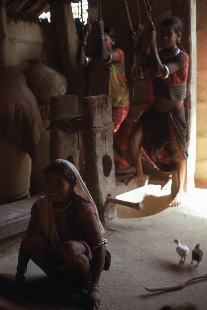 Three women husk rice inside of a building. Two women pull on rope over their heads as another crouches in the foreground.