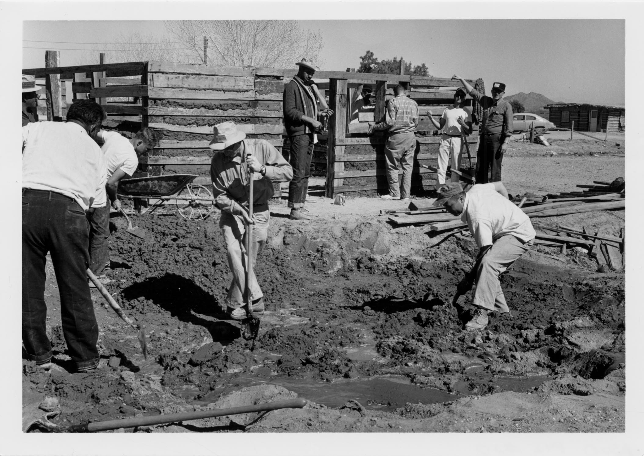 Black and white photo of three men dig in dirt with shovels. Another group of men stand along a wooden fence in the background.